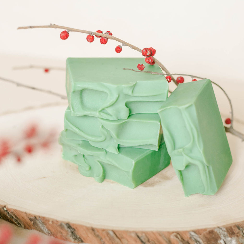 Two green handmade soap bars on a white surface with red berries and a branch.