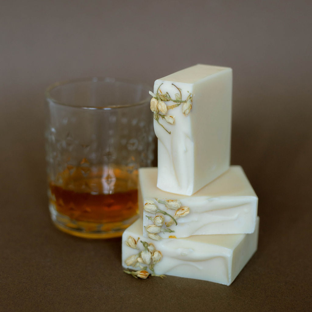 Three bars of white soap with floral decorations next to a glass of amber liquid on a brown surface.