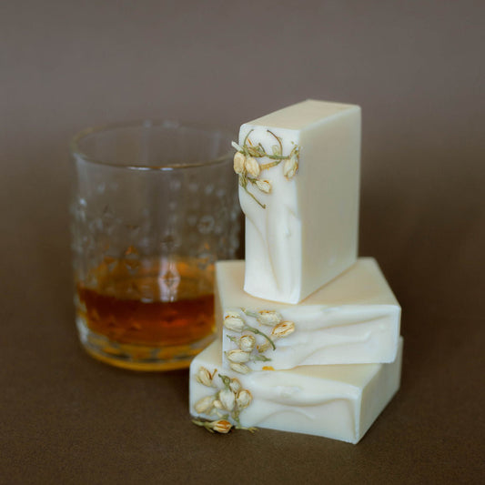 Three bars of white soap with floral decorations next to a glass of amber liquid on a brown surface.